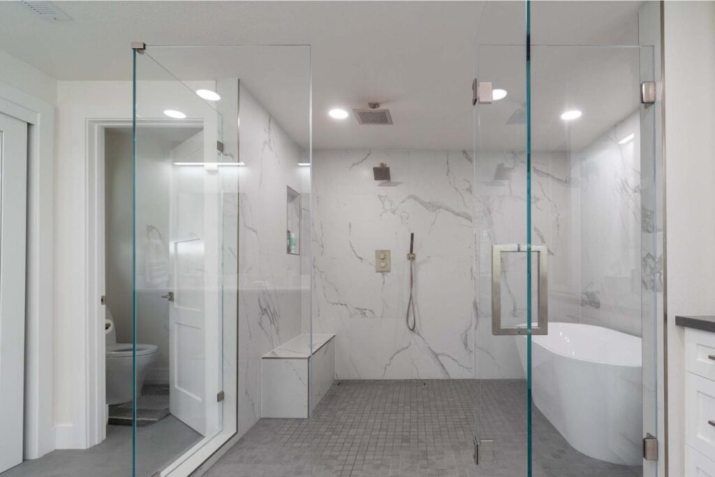 Sleek bath and shower remodel embraces minimalist aesthetic with floor-to-ceiling frameless glass enclosure, elegant freestanding tub, and white porcelain slabs.