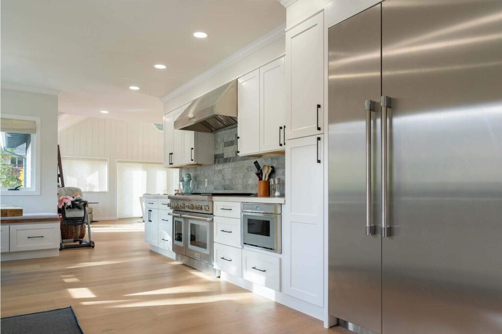 Galley-style white kitchen with professional-grade stainless steel refrigerator, light wood floors, and vaulted ceiling with natural light