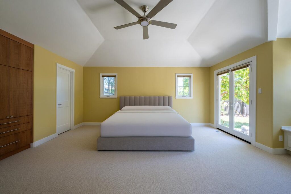 Bright second story addition bedroom with cost-effective layout featuring warm yellow walls, ceiling fan, multiple windows for natural light, and traditional design perfect for family expansion.